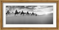 Tourists riding camels through the Sahara Desert landscape led by a Berber man, Morocco (black and white) Fine Art Print